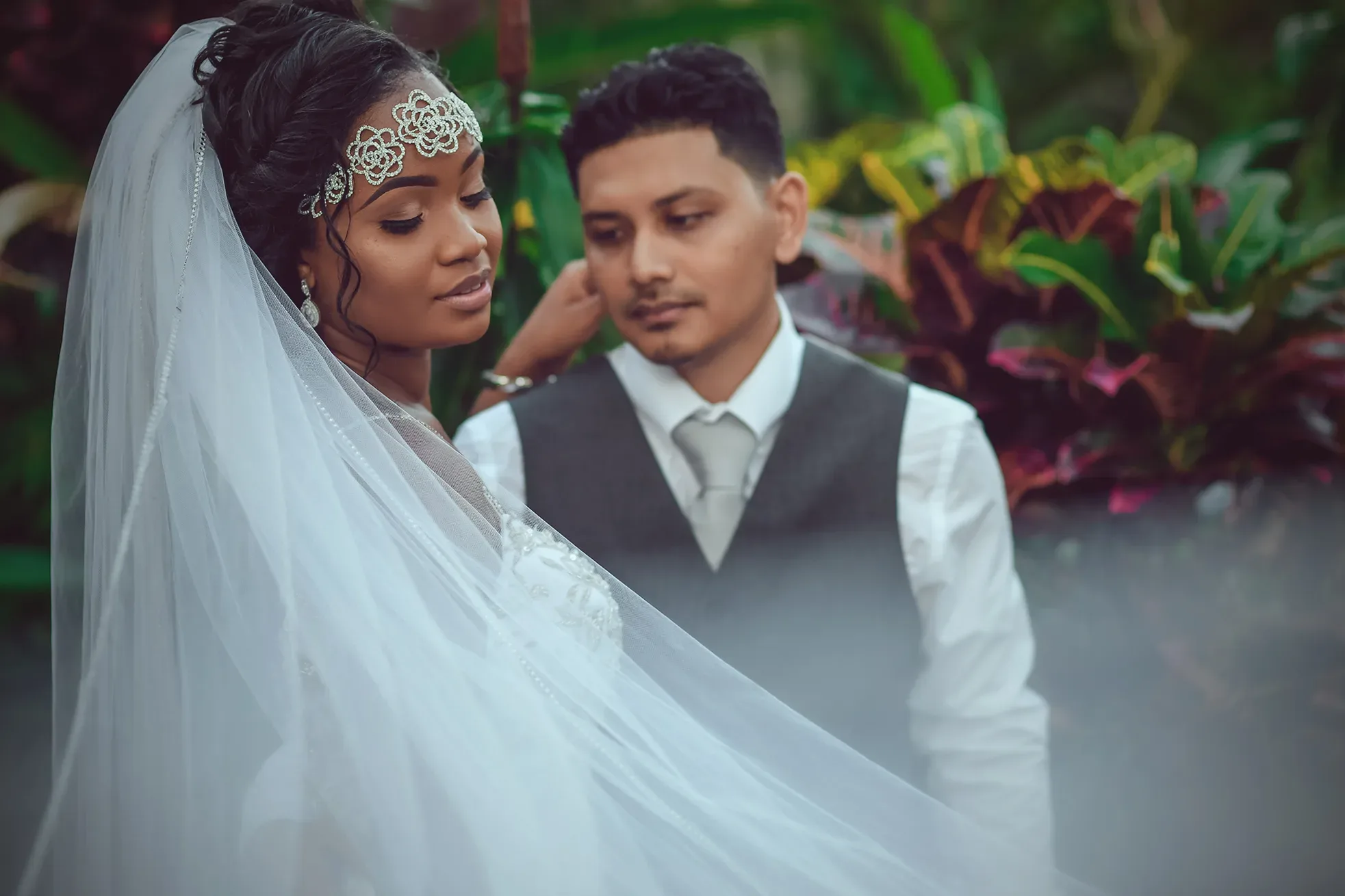 A bride with a crystal headband and veil looking down while her groom stands closely behind her in a lush garden.