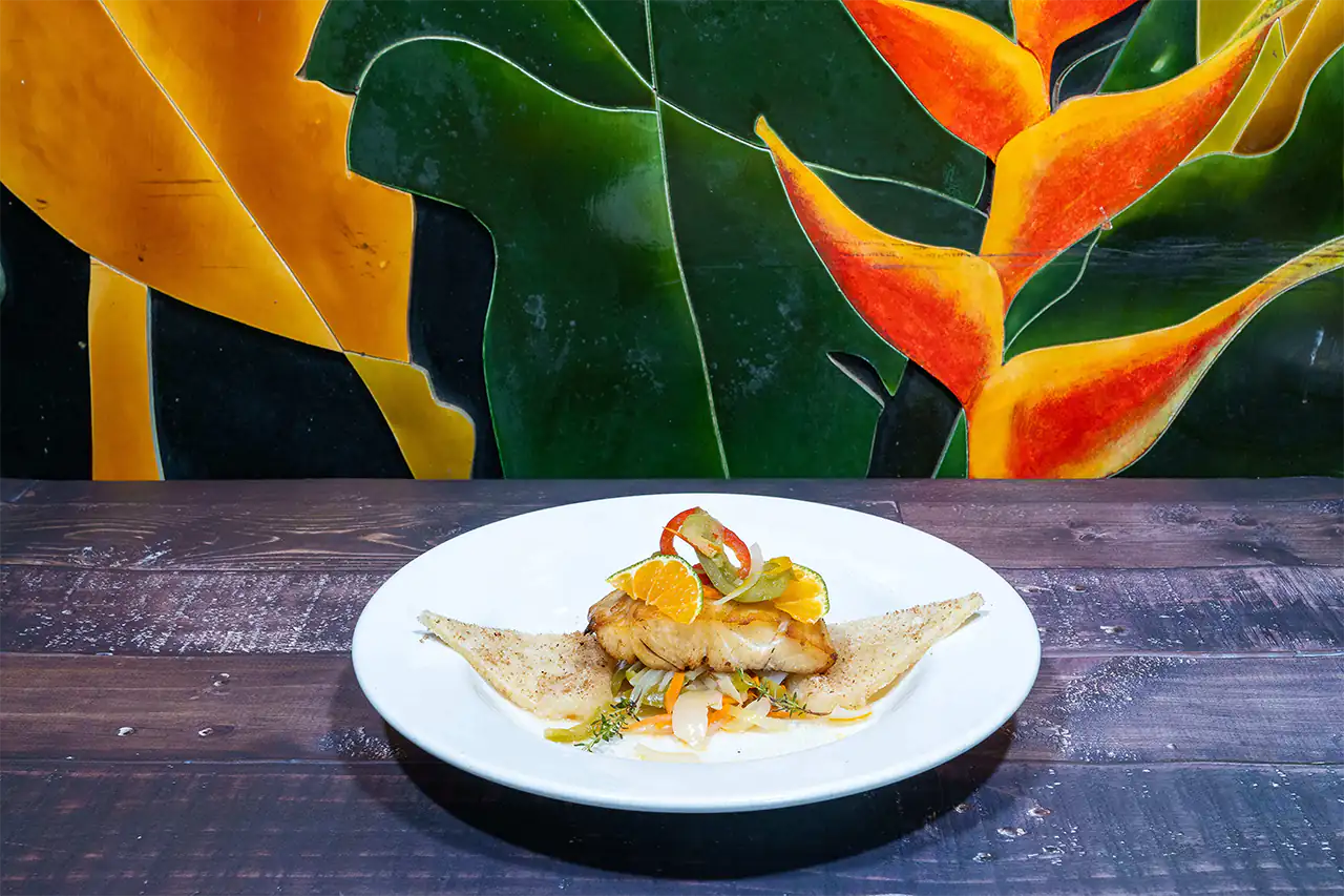 A plated Jamaican escovitch fish fillet topped with pickled vegetables, citrus slices, and scotch bonnet peppers, served between two triangles of fried bammy on a white plate against a tropical mural background.