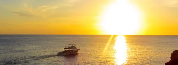 A small boat cruising on the ocean at sunset with a bright yellow sun reflecting on the water.